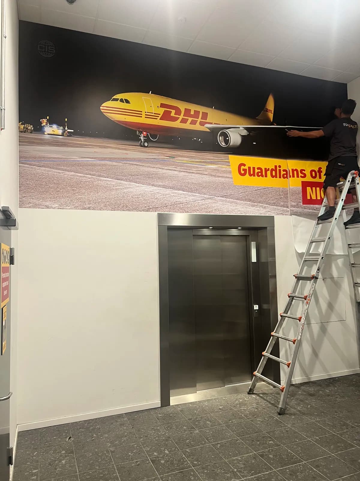 Montage af vægfolie med DHL-flymotiv, hvor montøren arbejder fra stige i trappeopgang