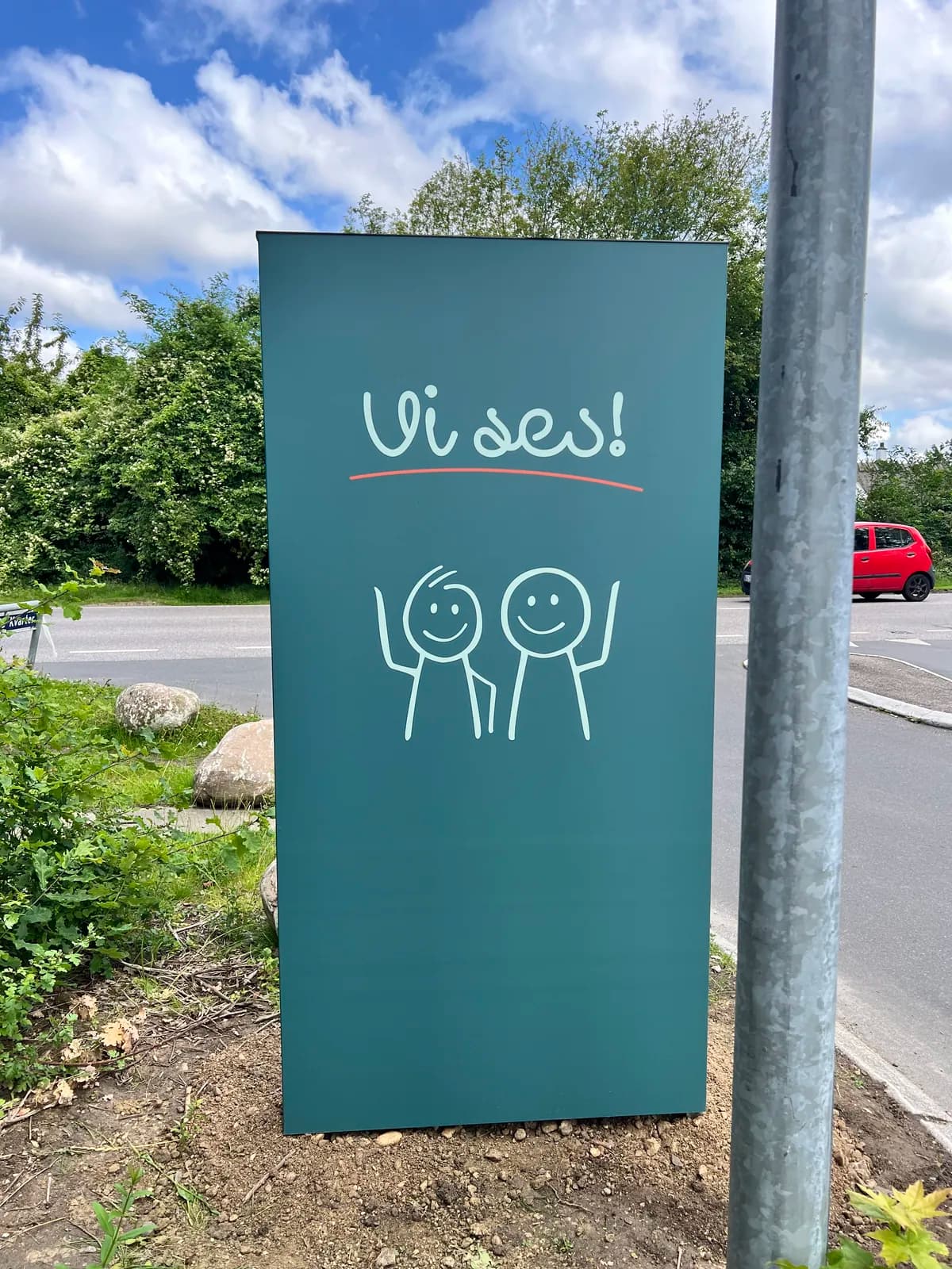 Fritstående grønt skilt med håndtegnet 'Vi ses!'-motiv ved udkørsel fra parkeringsområde