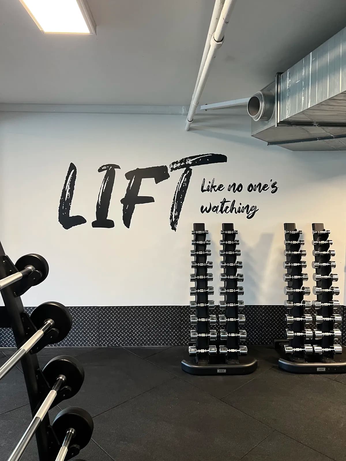 Vægfolie med teksten 'Lift like no one's watching' over håndvægtsstativer i fitnesscenter