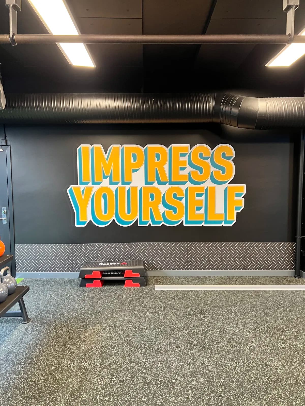 Vægfolie med teksten 'Impress yourself' i gult og petroleum på sort væg i fitnesscenter